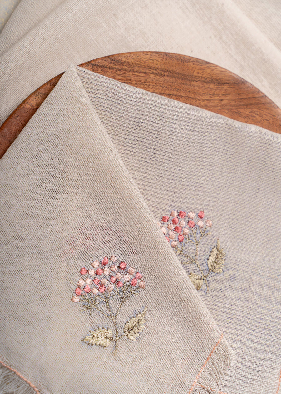 The Misty Garden Napkin brings delicate charm to the dining table with its soft, airy weave and intricate floral embroidery. Hand-stitched blossoms in warm pink tones, complemented by muted green leaves, add a subtle yet elegant touch. Finished with fine fringed edges, this napkin embodies effortless sophistication.