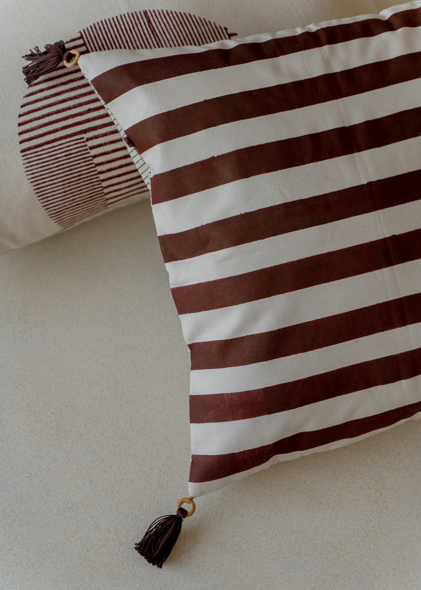 Ember Glow Cushion Cover – A striking cotton cushion cover featuring bold, hand-block-printed stripes in warm earthy tones. Finished with elegant tassels and wooden bead detailing, this artisanal piece adds depth and texture to any space, celebrating traditional craftsmanship with a modern aesthetic.