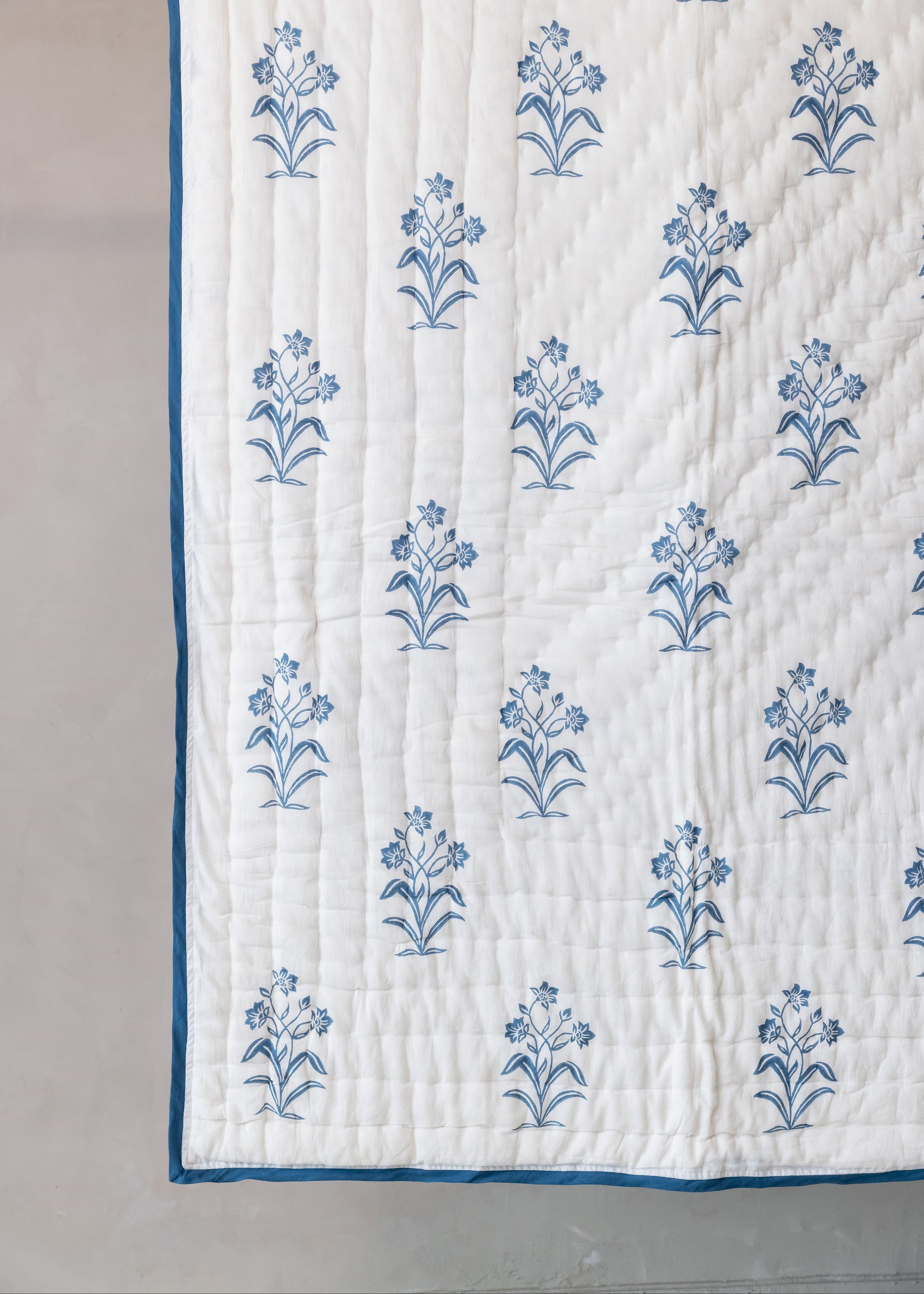 The Indigo Reverie Quilt, crafted from handwoven cotton, showcases a delicate floral pattern meticulously hand block printed in a serene indigo hue. Its soft, quilted texture and refined border add a timeless elegance, making it a perfect addition to any tranquil and inviting space.