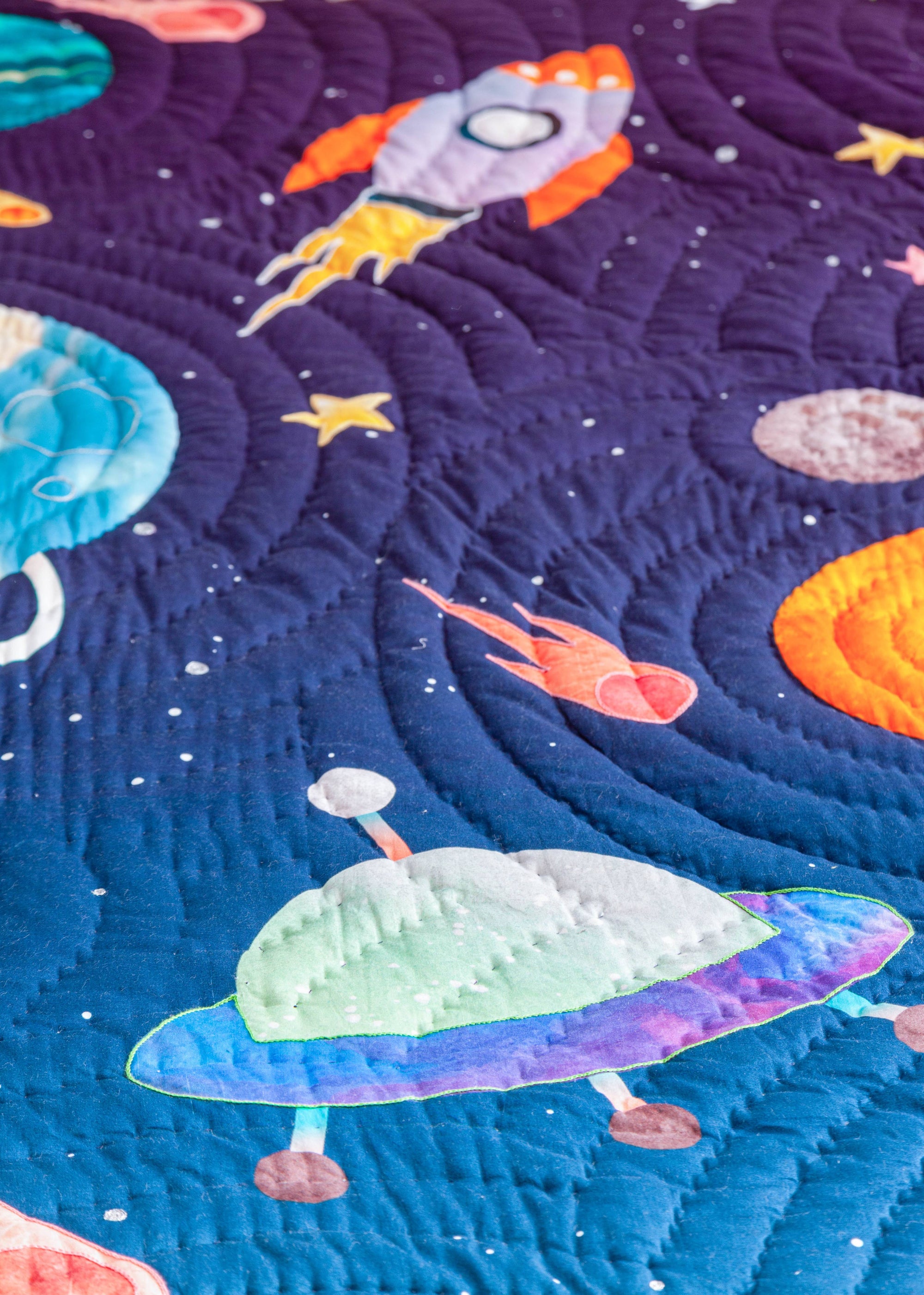 A close-up of the &quot;Tim, the Traveler Quilt,&quot; a handcrafted space-themed quilt featuring an appliqué design of a vibrant blue and green planet, a fiery comet, and scattered stars on a deep purple background. The quilt is folded to reveal its playful geometric patterned reverse side in shades of blue, purple, and orange. Placed on a soft white textured surface, an astronaut figurine lies beside it, enhancing the celestial theme.