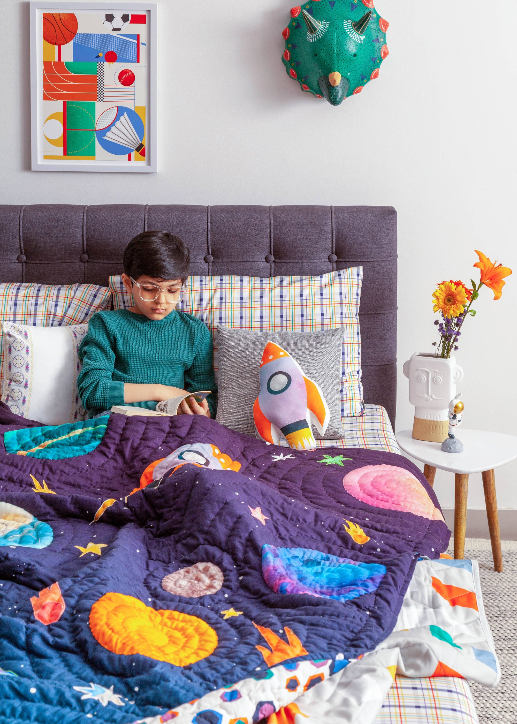 A cozy and imaginative bedroom scene featuring a young boy reading under the &quot;Tim, the Traveler Quilt.&quot; The quilt, handcrafted with intricate space-themed appliqué, showcases planets, rockets, and stars in vibrant colors. A matching rocket-shaped cushion adds to the celestial theme, while plaid pillows and a dark upholstered headboard provide a modern contrast. A side table with fresh flowers, a quirky astronaut figurine, and a framed sports-inspired artwork complete the playful yet stylish setting.