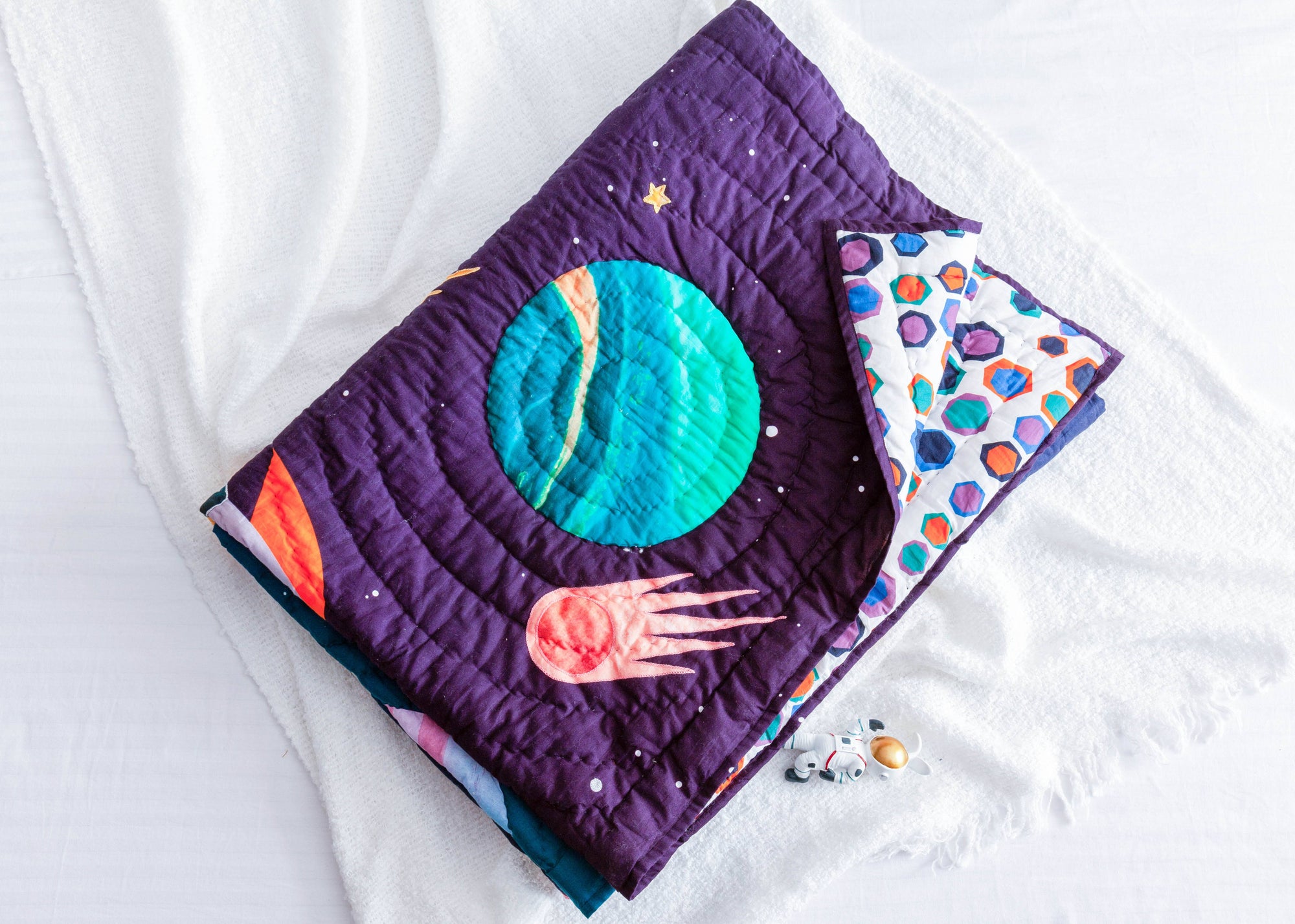 A close-up of the &quot;Tim, the Traveler Quilt,&quot; a handcrafted space-themed quilt featuring an appliqué design of a vibrant blue and green planet, a fiery comet, and scattered stars on a deep purple background. The quilt is folded to reveal its playful geometric patterned reverse side in shades of blue, purple, and orange. Placed on a soft white textured surface, an astronaut figurine lies beside it, enhancing the celestial theme.