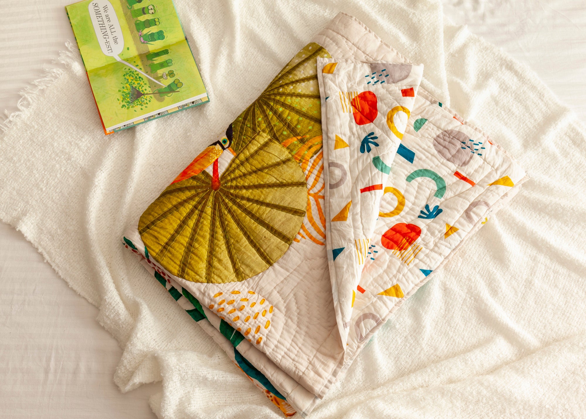 A folded The Forest is My Playground quilt, handcrafted from soft cotton, featuring a vibrant jungle scene with a toucan perched under large tropical leaves. The quilt’s reverse side displays a playful abstract pattern in bright, modern colors. Set against a cozy white blanket with a children&#39;s book nearby, the image captures warmth, comfort, and storytelling through textile artistry.