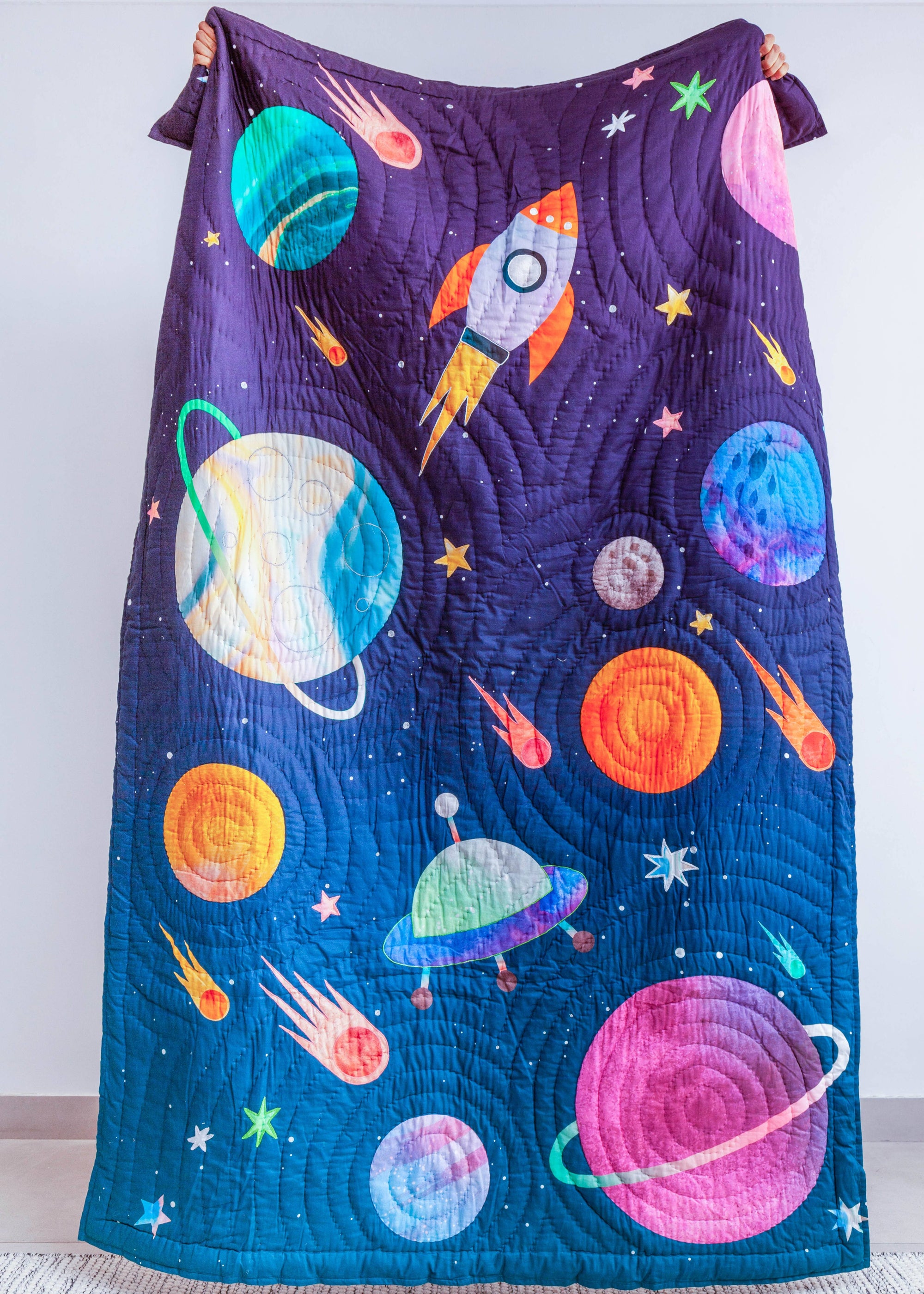 A cozy and imaginative bedroom scene featuring a young boy reading under the &quot;Tim, the Traveler Quilt.&quot; The quilt, handcrafted with intricate space-themed appliqué, showcases planets, rockets, and stars in vibrant colors. A matching rocket-shaped cushion adds to the celestial theme, while plaid pillows and a dark upholstered headboard provide a modern contrast. A side table with fresh flowers, a quirky astronaut figurine, and a framed sports-inspired artwork complete the playful yet stylish setting.