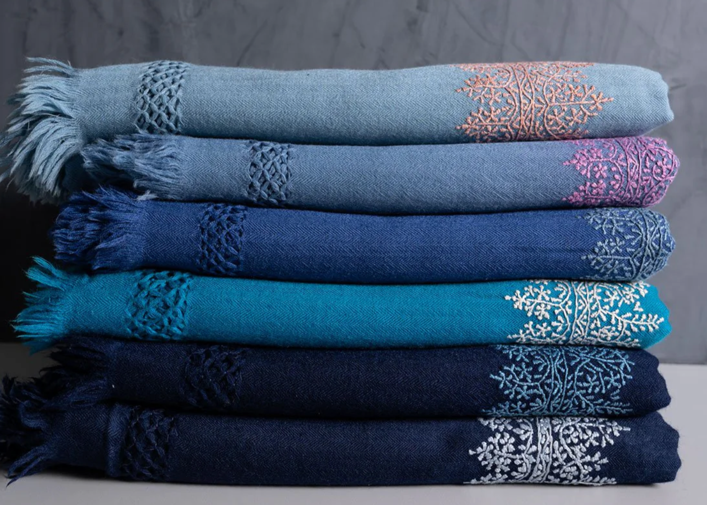 A neatly stacked selection of Chambi Bedspreads with Snow Shades, each in varying shades of blue, adorned with intricate embroidery and delicate fringe details. The gradient of hues, from sky blue to deep navy, showcases the fine craftsmanship and timeless elegance of these handcrafted textiles.