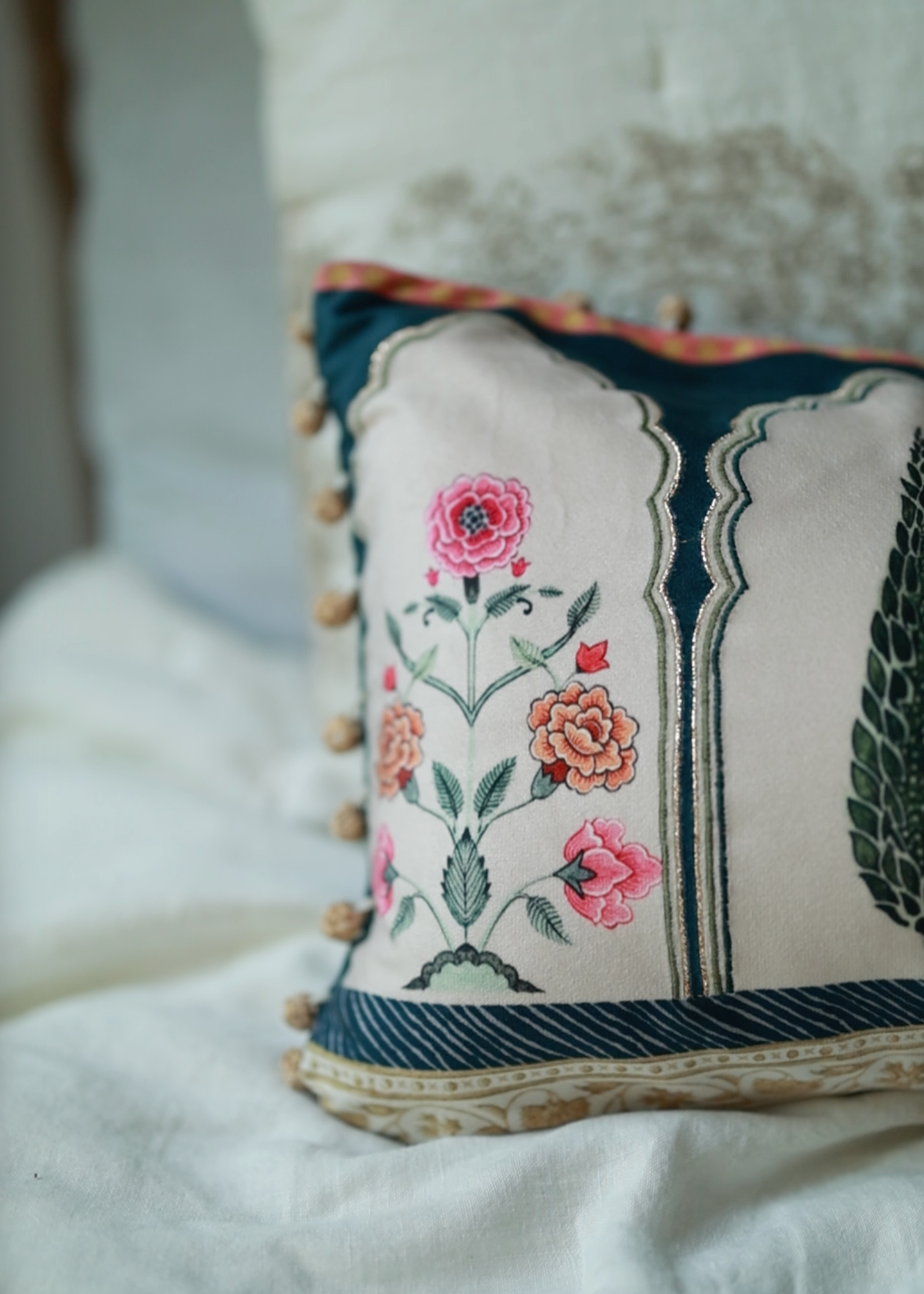 Garden Blossom Cushion Cover – This handcrafted cotton cushion cover features intricate floral embroidery inspired by traditional Mughal motifs. The delicate pink and orange blossoms contrast elegantly against a soft cream background, framed with deep blue detailing and finished with playful tassel accents.