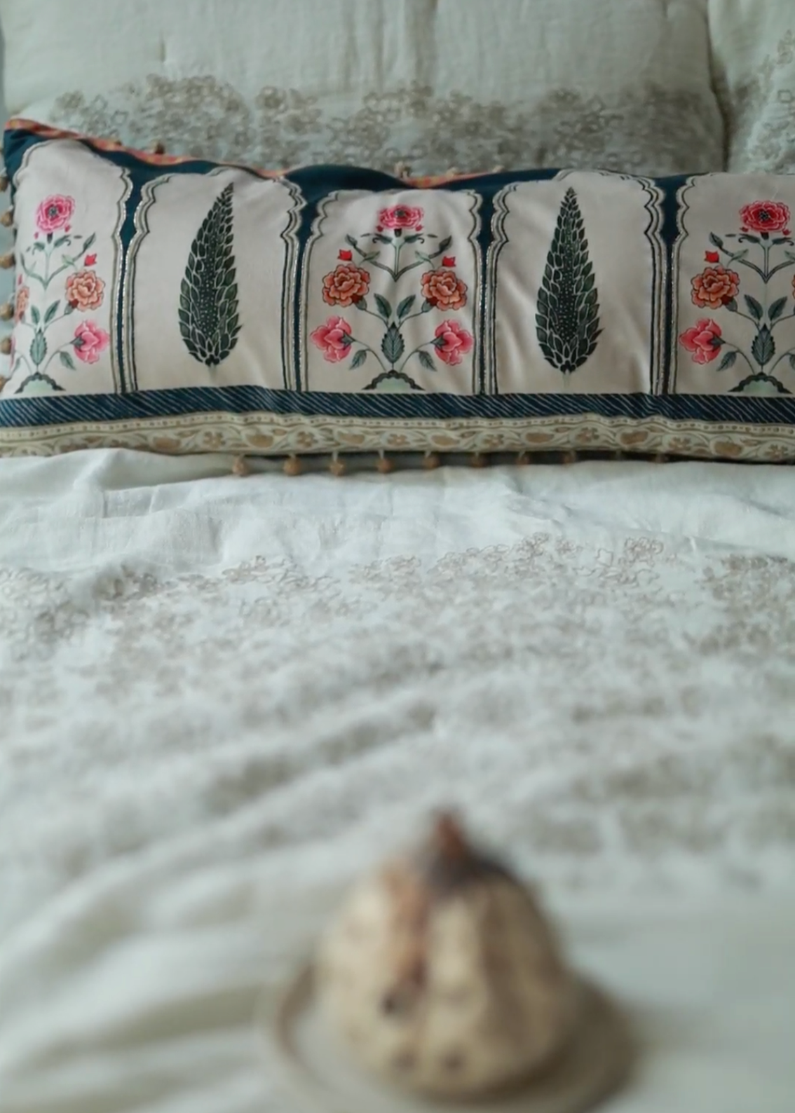 Garden Blossom Cushion Cover – This handcrafted cotton cushion cover features intricate floral embroidery inspired by traditional Mughal motifs. The delicate pink and orange blossoms contrast elegantly against a soft cream background, framed with deep blue detailing and finished with playful tassel accents.