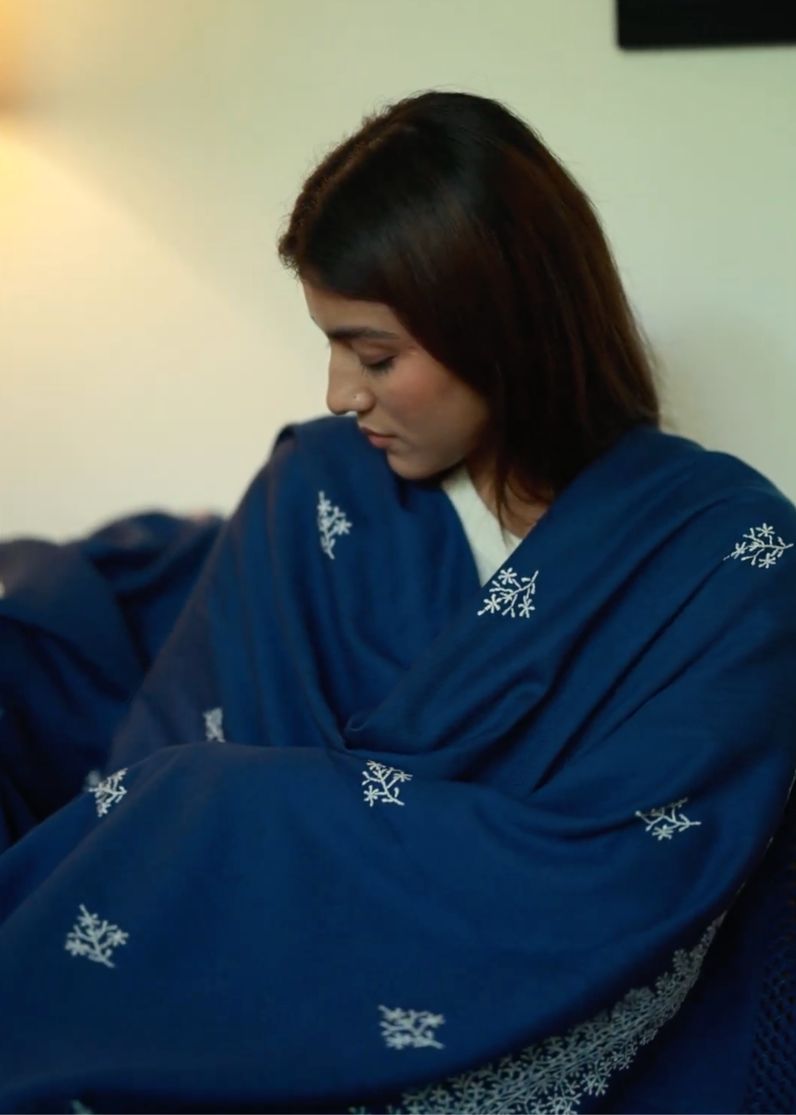 A woman wrapped in the Chambi Bedspread with Snow Shades, a deep blue textile adorned with delicate white embroidery. The soft fabric drapes around her, evoking warmth and comfort in a serene indoor setting.