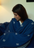 A woman wrapped in the Chambi Bedspread with Snow Shades, a deep blue textile adorned with delicate white embroidery. The soft fabric drapes around her, evoking warmth and comfort in a serene indoor setting.
